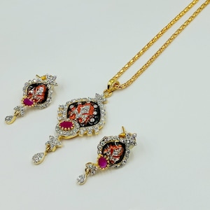 Puede incluir: Un conjunto de joyería en tono dorado que incluye un collar y pendientes a juego. Cada pieza presenta un diseño central de esmalte naranja y negro con cristales transparentes y una piedra rosa ovalada, rematado con un colgante de cristal.