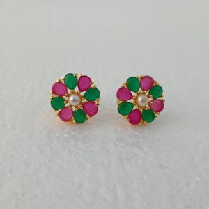 May include: A pair of gold-toned stud earrings. Each earring features a floral design with alternating pink and green teardrop-shaped gemstones surrounding a central pearl. The earrings are set against a white background.