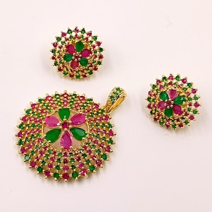 Puede incluir: Un conjunto de joyería compuesto por un colgante circular y dos pendientes a juego. Cada pieza presenta una base de metal dorado adornada con pequeñas piedras redondas de color verde vibrante y rosa en un patrón floral.