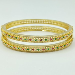 May include: Two gold bangles with a decorative pattern of small red, green, and white gemstones. The bangles have a textured design along the edges and a polished finish, suitable for jewelry.
