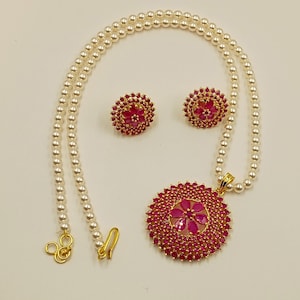 Puede incluir: Un conjunto de joyería compuesto por un collar y dos pendientes. El collar presenta una hilera de perlas blancas con un gran colgante redondo de tono dorado engastado con numerosas piedras rosas pequeñas y un racimo central en forma de flor.