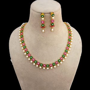 Puede incluir: Un conjunto de collar y pendientes dorados. El collar y los pendientes presentan un diseño floral con detalles verdes y rosas, intercalados con pequeñas perlas blancas. La joyería se exhibe en un maniquí.