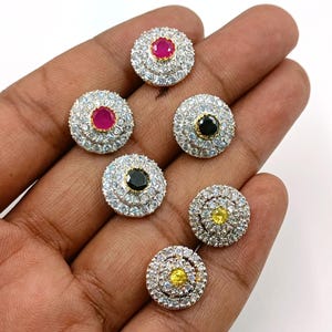 Puede incluir: Seis pendientes circulares de tipo botón con una piedra central rodeada por dos anillos concéntricos de pequeñas piedras claras. El conjunto incluye dos piedras rosas, dos negras y dos amarillas, montadas en un marco metálico con acabado pulido.