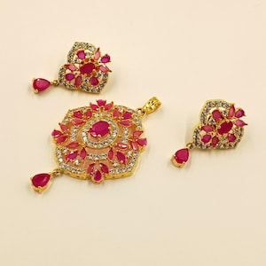 May include: A gold jewelry set consisting of a pendant and two matching earrings. Each piece features a central oval-cut pink stone surrounded by smaller pink marquise-cut stones and clear white crystals in a floral-inspired arrangement.