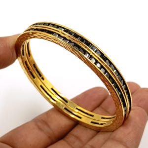 Könnte beinhalten: Ein goldfarbener Armreif mit einer texturierten, gehämmerten Außenfläche und einem zentralen Kanal, der mit einer durchgehenden Reihe kleiner, quadratisch geschliffener schwarzer Steine besetzt ist. Das Design besteht aus zwei parallelen Goldbändern.