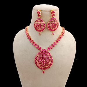 Puede incluir: Un conjunto de joyas de tono dorado con un collar y pendientes a juego. El collar tiene un colgante grande y redondo y una cadena de eslabones más pequeños en forma de hoja. Los pendientes tienen forma de lágrima. Ambas piezas están adornadas con vibrantes piedras preciosas de color rojo rubí.