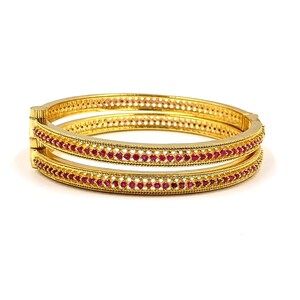 May include: Two gold-colored bangles with a repeating pattern of small, red gemstones. The bangles have a detailed design with a series of small holes along the edges. The jewelry is set against a white background.