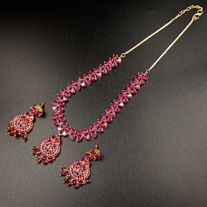 Op de afbeelding: Een goudkleurige sieradenset met een ketting en bijpassende oorbellen. De ketting en oorbellen zijn versierd met levendige, robijnkleurige edelstenen. De oorbellen hebben een cirkelvormig ontwerp met bungelende robijnaccenten. De ketting heeft een bloemenpatroon.