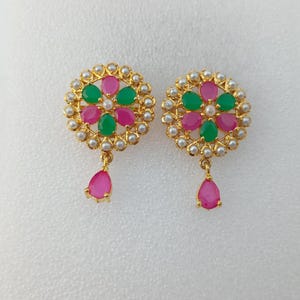 Può includere: Un paio di orecchini color oro. Ogni orecchino presenta un design floreale con pietre preziose rosa e verdi, circondate da piccole perle bianche. Una gemma rosa a forma di goccia pende dalla parte inferiore di ogni orecchino.
