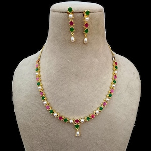 Op de afbeelding: Een goudkleurige ketting- en oorbellenset. De ketting en oorbellen hebben een herhalend patroon van groene, roze en rode edelstenen, afgewisseld met witte parels. De oorbellen hebben een druppelvormige parel.