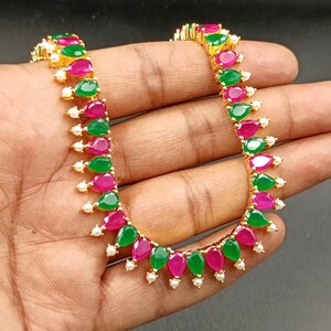 May include: A gold-toned necklace featuring alternating green and magenta teardrop-shaped gemstones. Small white pearls are interspersed between the gemstones. The necklace is held in a hand against a dark background.