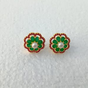 May include: A pair of gold-toned flower-shaped stud earrings. Each earring features a central pearl surrounded by green gemstones, with a border of red gemstones. The earrings are set against a white background.