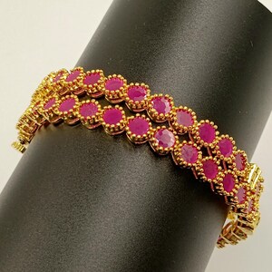 Op de afbeelding: Een goudkleurige armband met twee parallelle rijen ronde, fel magenta-roze stenen. Elke steen is afzonderlijk gezet in een getextureerd, kraalachtig goudkleurig metalen frame, wat zorgt voor een doorlopend, geschakeld ontwerp rond de houder.