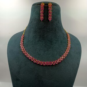 Peut inclure: Un collier doré et une paire de boucles d'oreilles assorties, ornés de petites pierres fuchsia taille marquise disposées en plusieurs rangées. Le collier présente une structure en chaîne souple et les boucles d'oreilles sont allongées.