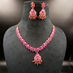 Puede incluir: Un conjunto de collar y pendientes dorados con piedras preciosas rosas vibrantes. El collar tiene un diseño floral con un colgante, y los pendientes son colgantes florales a juego. Las joyas se exhiben en un maniquí negro.