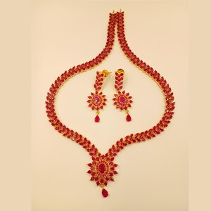 May include: A gold-coloured jewellery set comprising a necklace and matching earrings. The necklace and earrings are embellished with bright red gemstones. The necklace has a central floral design with a teardrop pendant. The earrings have a floral design with a teardrop pendant.