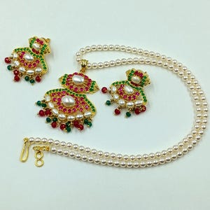 May include: A jewelry set consisting of a pearl necklace with a gold-tone pendant, and two matching earrings. Each piece features white pearls, vibrant red stones, and small green beads set in a gold-colored metal frame with a hook clasp.