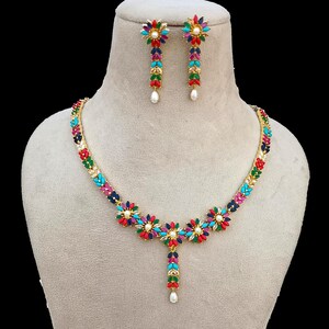 Puede incluir: Un conjunto de collar y pendientes dorados. El collar y los pendientes presentan diseños florales coloridos con detalles en rojo, azul, verde y turquesa. Cada pendiente y el colgante del collar terminan con una perla blanca.