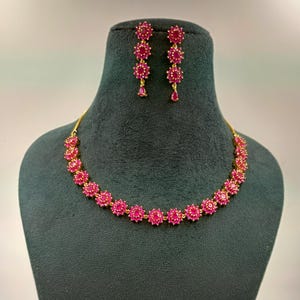 May include: A gold-toned necklace and matching drop earring set featuring vibrant pink, flower-shaped stones. The necklace consists of a single row of these floral clusters, while each earring features a vertical arrangement of three matching floral stones.