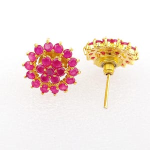 May include: A pair of gold-colored stud earrings. Each earring features a floral design composed of numerous small, round, magenta-colored gemstones. The earrings are set against a white background, highlighting the jewelry's details.