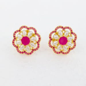Puede incluir: Un par de pendientes de botón dorados con forma de flor. Cada pendiente presenta una piedra central de color fucsia rodeada de acentos blancos con forma de perla y un borde de pequeñas piedras preciosas rojas. Sobre un fondo blanco.
