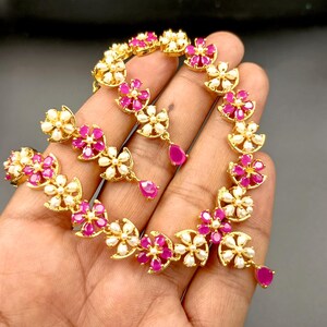 Op de afbeelding: Een goudkleurige ketting- en oorbellenset met een bloemmotief. De bloemen hebben roze centra en witte bloemblaadjes, met een druppelvormige roze edelsteen die aan elke oorbel hangt. De sieraden worden tegen een donkere achtergrond gepresenteerd.