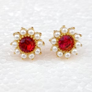 May include: A pair of gold-coloured stud earrings. Each earring has a central, round, red gemstone surrounded by small, white pearls. The pearls are arranged in a flower-like pattern around the red stone.