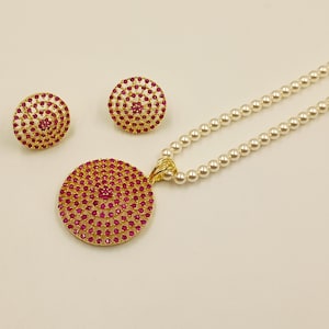 Puede incluir: Un conjunto de joyería compuesto por un collar y dos pendientes. El collar presenta una hilera de perlas blancas y un gran colgante circular de tono dorado incrustado con pequeñas piedras de color rosa vibrante en círculos concéntricos.