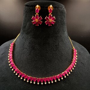 May include: A gold-coloured necklace and earring set. The necklace features a series of teardrop-shaped, ruby-coloured stones with small white accents. The matching earrings are flower-shaped, with ruby-coloured petals and gold-coloured centres. The jewellery is displayed on a black mannequin.