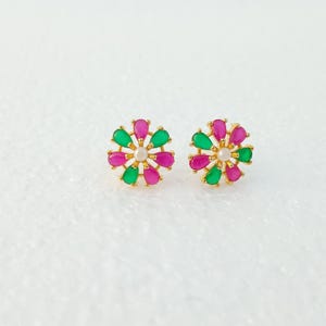 Può includere: Un paio di orecchini a bottone dorati. Ogni orecchino presenta un design floreale con pietre a forma di goccia verdi e rosa alternate, che circondano una perla bianca centrale. Gli orecchini sono su uno sfondo bianco.