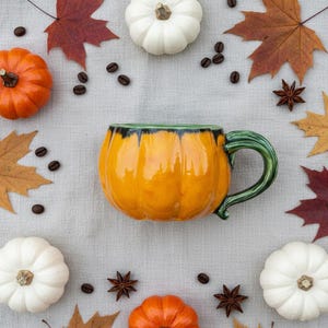 Könnte beinhalten: Eine orangefarbene, kürbisförmige Keramiktasse mit grünem Henkel und Rand. Umgeben von kleinen Kürbissen, Herbstblättern, Kaffeebohnen und Sternanis auf einer hellgrauen Oberfläche.
