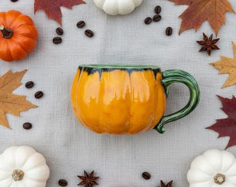 May include: An overhead shot of a pumpkin-shaped ceramic mug with a green handle and rim. The mug is surrounded by small pumpkins, fall leaves, coffee beans, and star anise on a light gray surface.