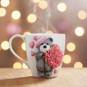 Könnte beinhalten: Weiße Keramiktasse mit einem dekorativen grauen Teddybären, der eine rosa-weiß gestreifte Mütze trägt und einen Strauß rosa Rosen hält. Zwei rosa Herzen sind auf der Tasse. Dampf steigt aus der Tasse auf.