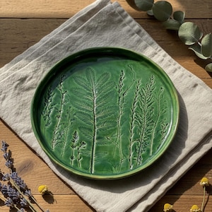 Puede incluir: Un plato redondo de cerámica verde con un diseño botánico en relieve que presenta varias plantas y hierbas. El plato está sobre una servilleta de lino de color neutro, con una superficie de madera en el fondo. Diámetro aproximado de 25 cm.