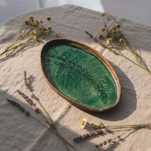 May include: An oval ceramic tray with a glossy emerald green glaze featuring a detailed fern leaf impression. The tray has a raised, unglazed brown rim that contrasts with the vibrant green center, creating a distinct botanical design element.