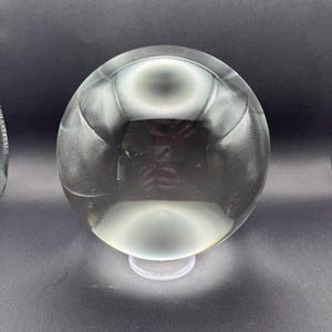 Op de afbeelding: Een heldere, bolvormige glazen bol die op een kleine, transparante ronde standaard rust. Het gladde, gepolijste oppervlak van de bol reflecteert de omgeving, wat zorgt voor vervormde cirkelvormige lichtpatronen en een reflectie van een camera.