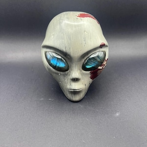 May include: A carved stone alien head sculpture featuring a smooth, light gray surface with dark gray veining. The eyes are inlaid with iridescent blue labradorite gemstones, and there are distinct dark red paint splatters on the forehead and cheek.