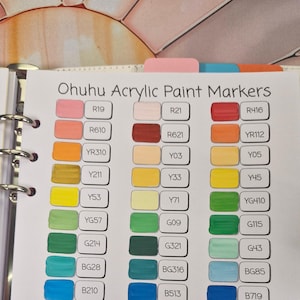 Op de afbeelding: Een kleurenkaart voor Ohuhu Acrylverf Markers. De kaart toont een verscheidenheid aan kleuren, elk gelabeld met een unieke code. De kleuren variëren van roze en oranje tot groen, blauw, paars en bruin. De kaart is georganiseerd in een ringband.