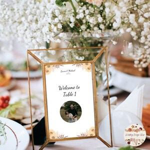Op de afbeelding: Een goudkleurig tafelbord met de tekst "Welkom bij Tafel 1" en een foto van een stel. Het bord is versierd met bloemaccenten en staat op een tafel met een bloemstuk op de achtergrond. Het bord is voor een bruiloft.