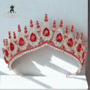 May include: A silver-toned metal tiara featuring numerous clear rhinestones and large, teardrop-shaped red crystals. The crown has a symmetrical design with multiple vertical points of varying heights, all encrusted with sparkling stones.
