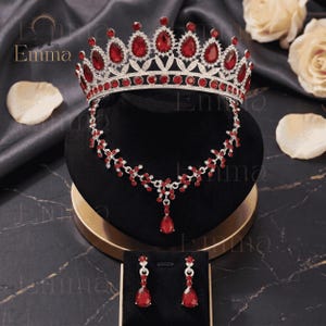 May include: A silver-toned jewelry set featuring a tiara, a necklace, and a pair of drop earrings. Each piece is adorned with deep red, pear-shaped crystals and clear rhinestones. The tiara has a high, arched design, while the necklace features a delicate vine-like pattern with a central teardrop pendant. The earrings match the teardrop design.