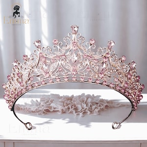 May include: A rose gold tiara featuring an intricate metal filigree design adorned with numerous light pink and clear crystals. The crown has a curved base with a row of pink stones and multiple upward-pointing peaks set with faceted gems.