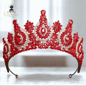 Op de afbeelding: Een metalen tiara met talloze gefacetteerde rode kristallen in een symmetrisch, sierlijk patroon. Het ontwerp bevat een verhoogde centrale piek, kleinere zijpieken en een gebogen basis, allemaal versierd met fonkelende rode stenen.