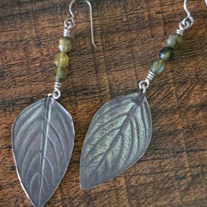 May include: A pair of silver leaf earrings with a textured finish. Each earring features a leaf-shaped pendant with detailed veining and a strand of green beads. The earrings have a hook closure.