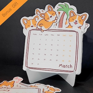 May include: A desk calendar featuring a March calendar page with corgi dog illustrations. The calendar has a white stand and is decorated with corgi stickers. The words "Bonus Gift" are in the top left corner.