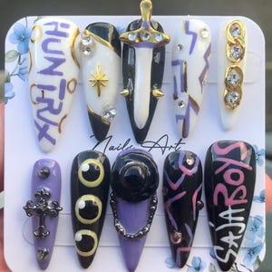 Puede incluir: Un juego de diez u&ntilde;as postizas en forma de estilete con una paleta de colores negro, blanco y morado. Los dise&ntilde;os incluyen el texto "HUNTRIX" y "SAJABOYS", un dije de espada en 3D, una cruz, pedrer&iacute;a, estrellas doradas y motivos oculares.