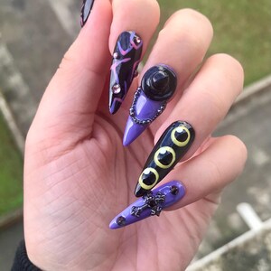 Puede incluir: U&ntilde;as postizas en forma de estilete con una paleta de colores morado y negro. Los dise&ntilde;os incluyen un sombrero de bruja negro, tres ojos amarillos con pupilas negras, una cadena plateada, una cruz negra y peque&ntilde;os brillantes rosas sobre base oscura.