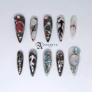 Puede incluir: Un juego de diez uñas postizas en forma de estilete con diseños en negro, blanco y rojo. Los adornos incluyen pedrería transparente y azul, grandes cabujones iridiscentes, peces koi pintados a mano y caracteres kanji japoneses. El logotipo Astreya Nails está centrado.