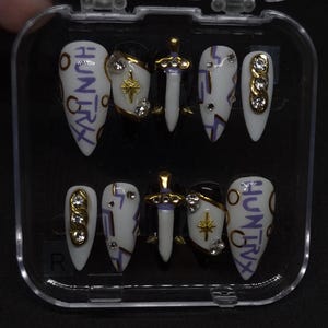 Puede incluir: Un juego de diez u&ntilde;as postizas en un estuche de pl&aacute;stico transparente. Las u&ntilde;as presentan una paleta de colores blanco y negro con letras moradas que dicen HUNTRIX, emblemas de espada dorados, detalles de estrellas y pedrer&iacute;a incrustada.