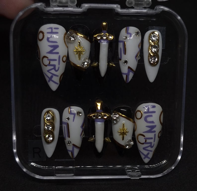 Puede incluir: Un juego de diez u&ntilde;as postizas en un estuche de pl&aacute;stico transparente. Las u&ntilde;as presentan una paleta de colores blanco y negro con letras moradas que dicen HUNTRIX, emblemas de espada dorados, detalles de estrellas y pedrer&iacute;a incrustada.